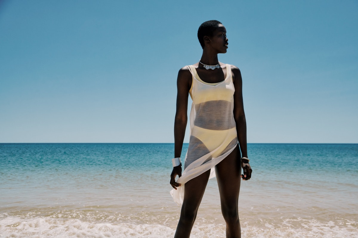 Woman in a white swimsuit standing on a beach with clear blue water and sky.