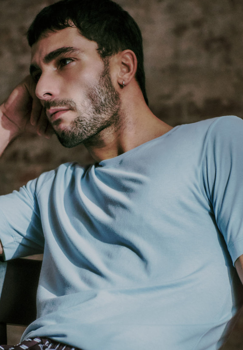 A man wearing a light blue crew-neck t-shirt, shown in a close-up portrait against a textured, warm-toned background.