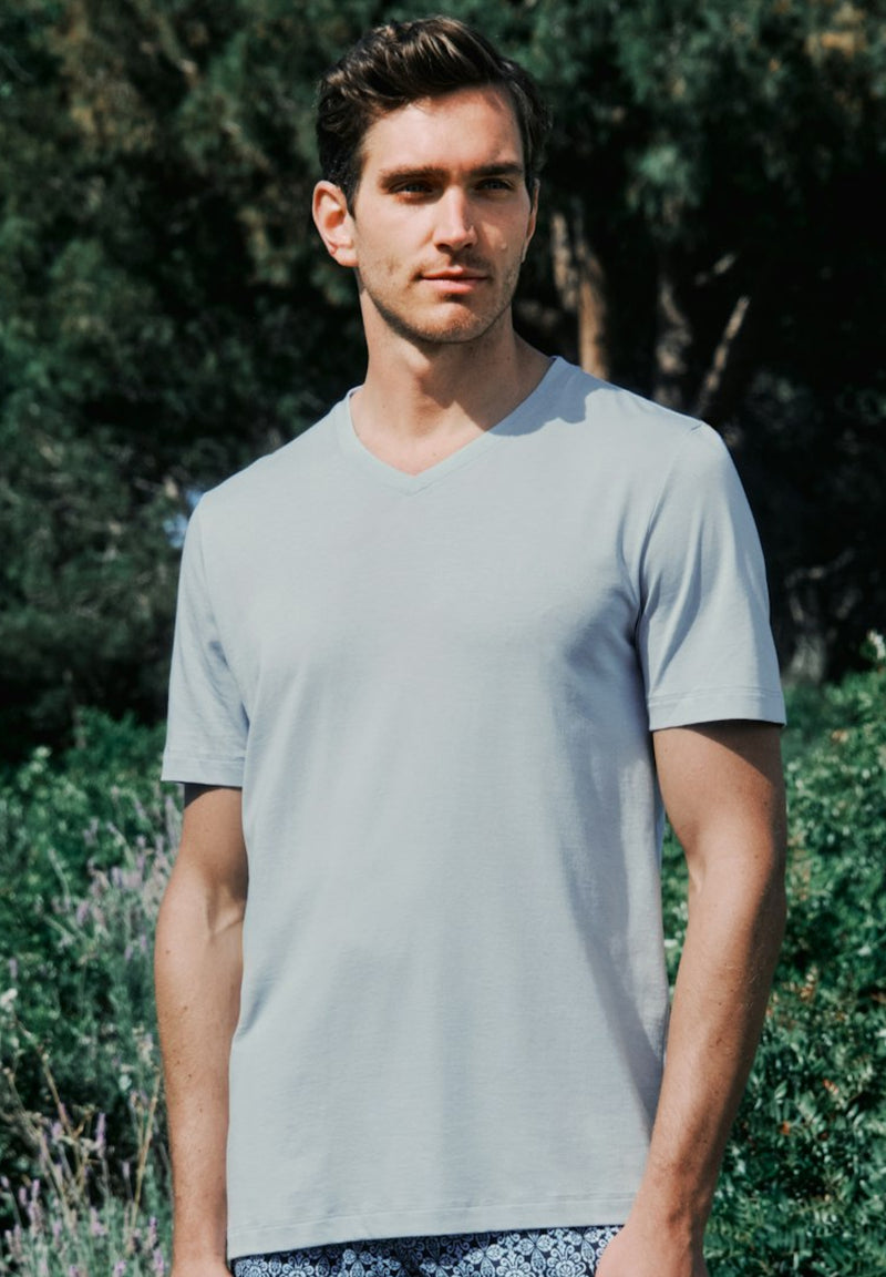 Man wearing a light gray t-shirt standing outdoors with greenery in the background