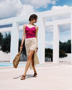 Woman in a pink top and beige skirt standing in front of a white architectural structure with blue sky and trees in the background.