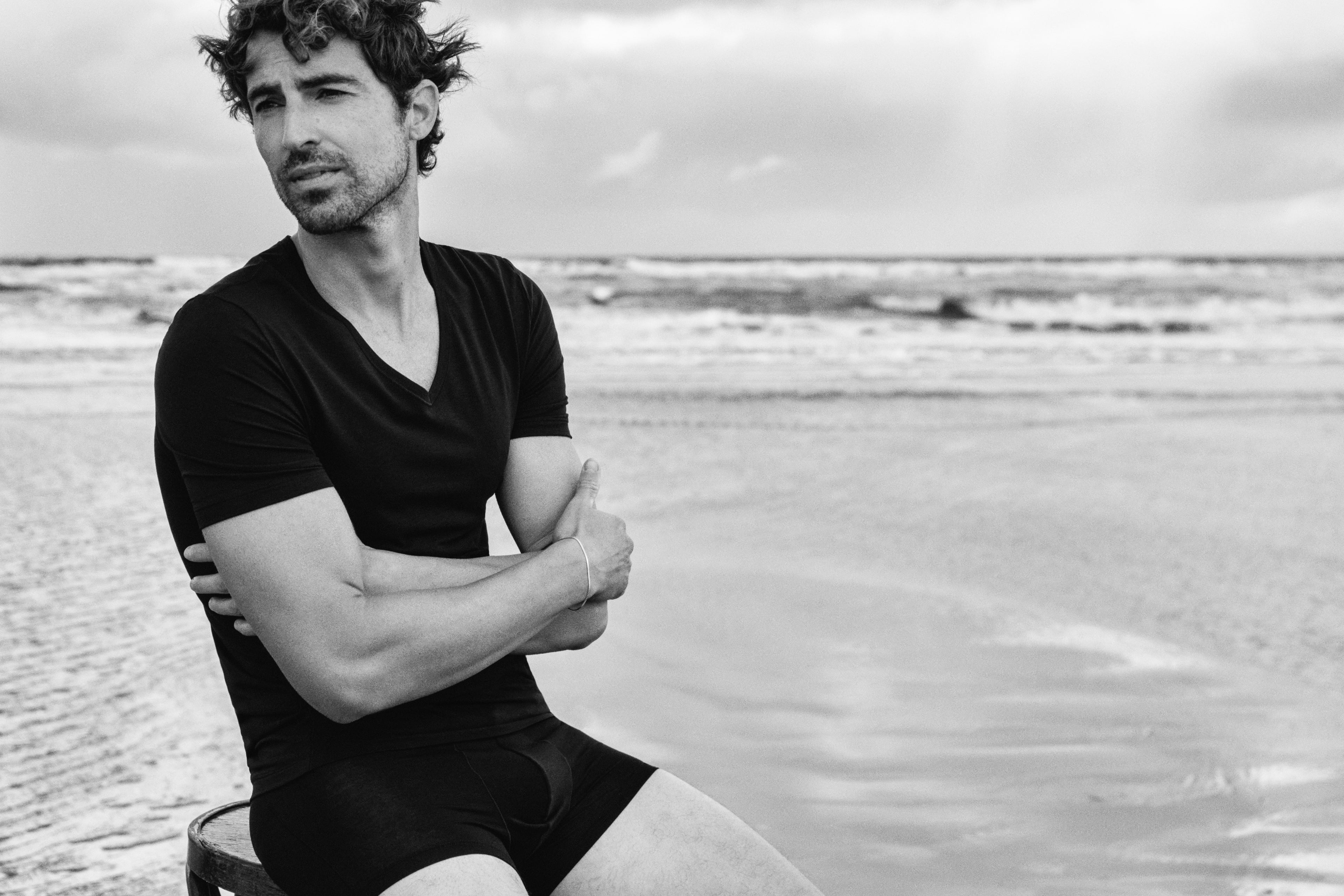 A man sitting on a beach wearing a black v-neck t-shirt and black boxer briefs, captured in a black and white photograph.